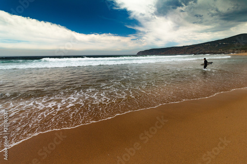 View of the Guincho beach near Atlantic coast. Surfer on the ocean coast in a wet suit with surfboard. Landscape of sunny day, blue sky and a mountain in distance. Cascais. Portugal.