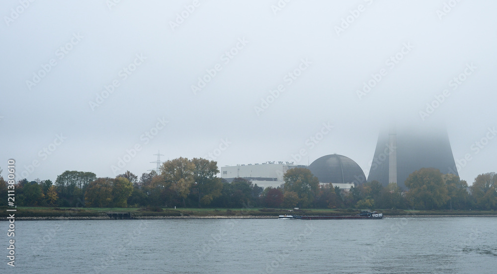 Fototapeta premium Foggy autumn morning in Germany on the river Rhine, a flowing barge. Visible chimney of a nuclear power plant in the fog.
