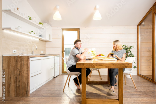 Young and happy couple having a breakfast sitting at the wooden table of the dining room of the modern country house. Wide angle view