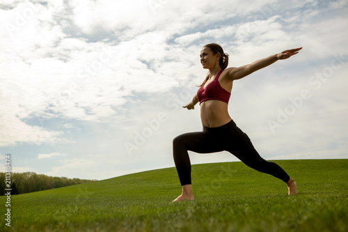 Smiling fit woman is exercising on lawn. She is training balance and flexibility while standing in lunge and stretching arms to sides. Work out outdoors for health concept