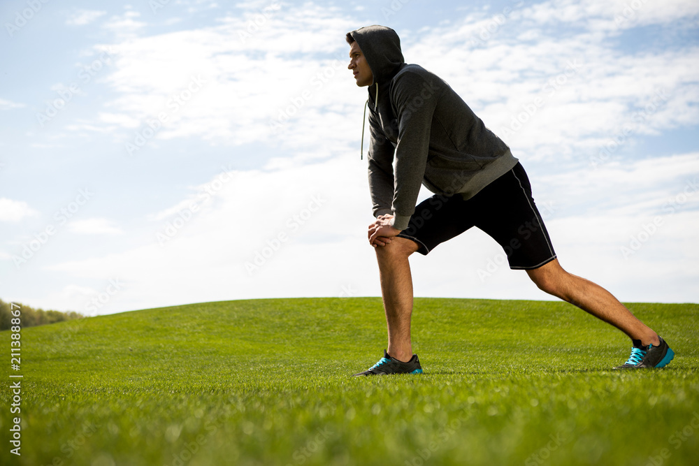 Concentrated man is doing lunges on green lawn during lovely day. He is stretching and warming-up on grass while looking ahead. Copy space in left side Concentrated man is doing lunges on green lawn during lovely day. He is stretching and warming-up on grass while looking ahead. Copy space in left side