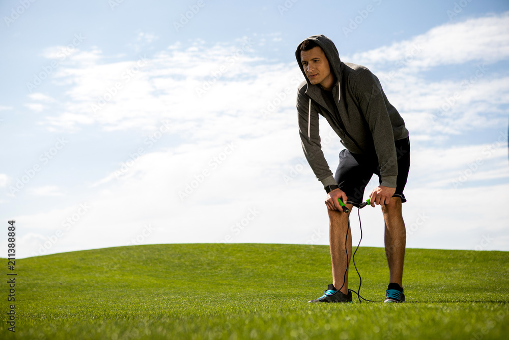 Calm man is having break after jumping with skipping rope on lawn. He is bending and holding equipment. Copy space in right side Calm man is having break after jumping with skipping rope on lawn. He is bending and holding equipment. Copy space in right side