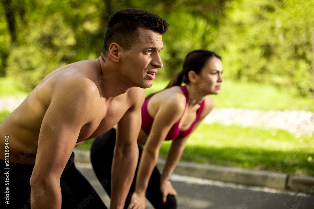 Two sportspeople are having break during cardio training in countryside. They are standing on road and bending body with hands on knee. Man and woman are feeling tired after exercising together Two sportspeople are having break during cardio training in countryside. They are standing on road and bending body with hands on knee. Man and woman are feeling tired after exercising together