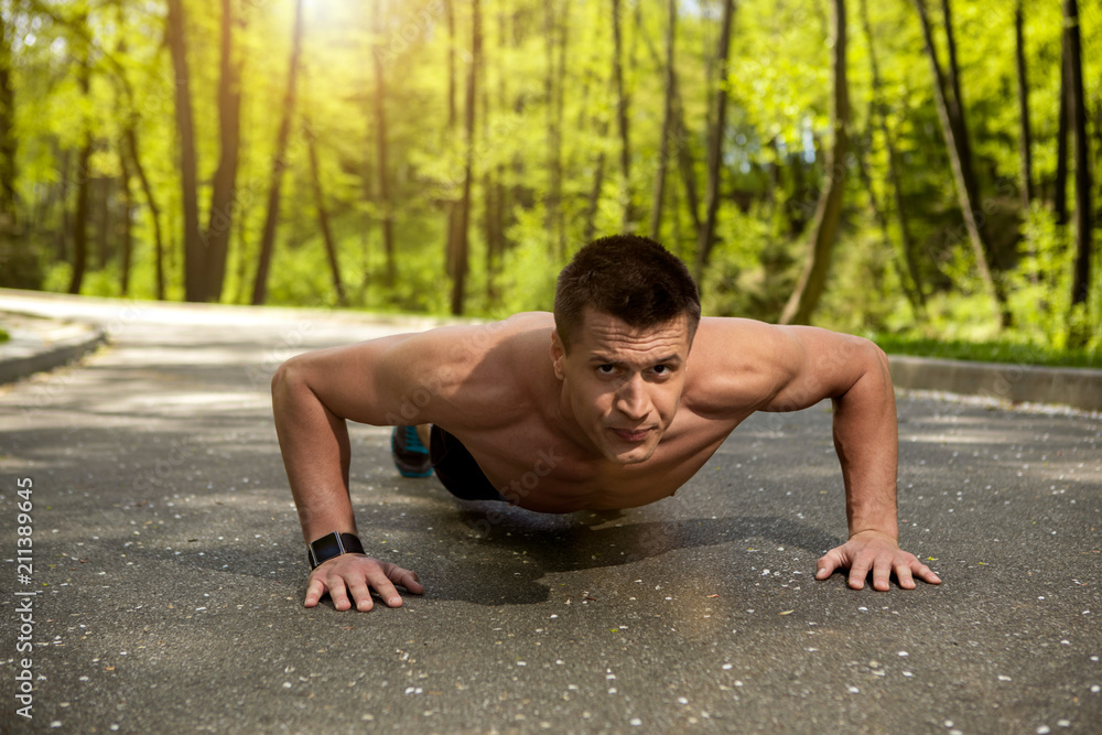 Fototapeta premium Athletic guy is concentrated on body workout. He is training pectorals with press-ups and wearing smartwatch. Male is exercising on road in green forest