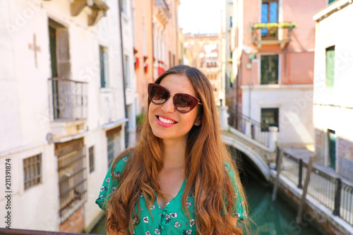 Smiling young woman with sunglasses and green dress in Venice. Happy beautiful girl standing on Venice bridge on canal, Italy
