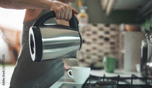 Close up focus on female hand holding kettle and pouring water into cup. Lady is preparing morning hot drink for breakfast in kitchen. Copy space in right side photo