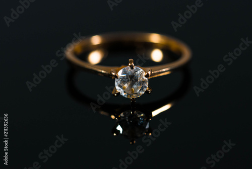 Real gold ring with diamond reflecting on black shiny surface close up macro shot.