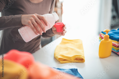 Close up of female hands pouring detergent into bottle lid. She is standing at white table with folded clothes and can of soap nearby