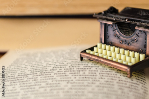 Small typewriter in copper tones over a page of a book. Decoration item. Concept of reading, writing, old times, past. Close up. Blurred background.