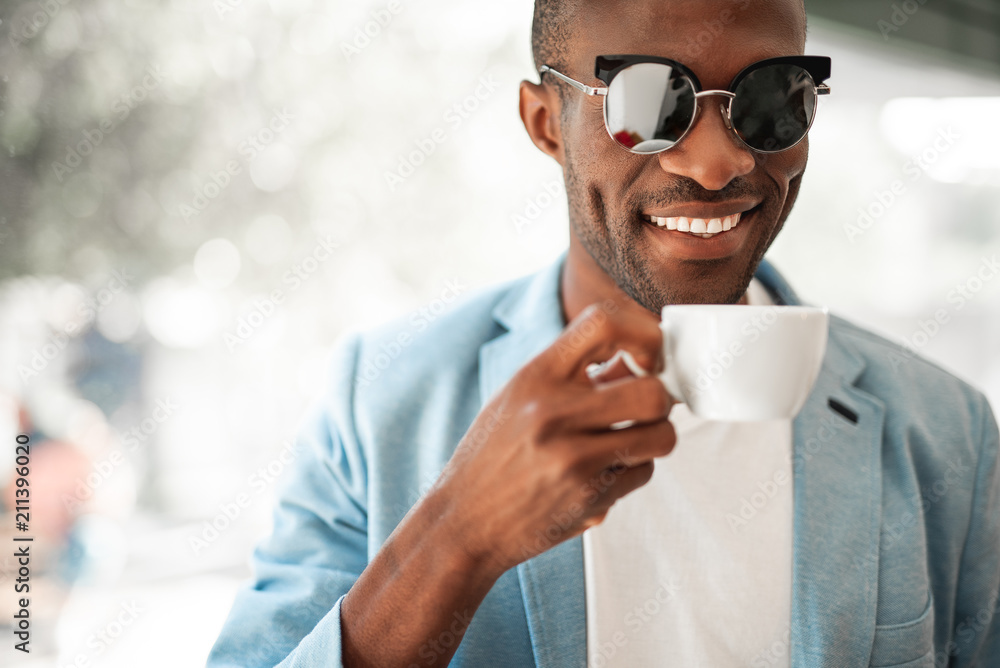 Positive man with attractive smile tasting mug of beverage during labor. He is wearing cool glasses Positive man with attractive smile tasting mug of beverage during labor. He is wearing cool glasses