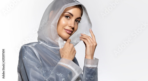 Portrait of cute sensual brunette in transparent raincoat posing at studio. Engaging girl with natural make-up looking at camera with tenderness. Isolated on grey background