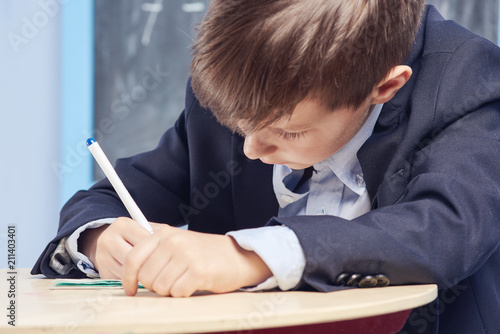Cheerful Caucasian schoolboy in uniform carefully doing task inclassroom. First time to school. Back to school. Close view.