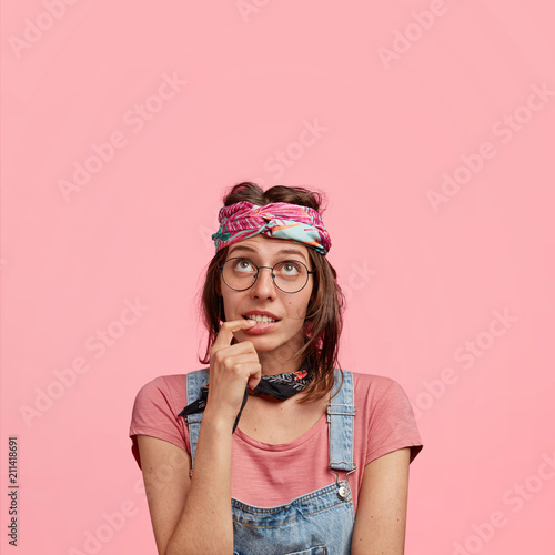 Pretty stylish female hippy in casual wear, has doubtful expression, focused upwards, holds fore finger in mouth, tries to find best solution, looks with indecisiveness, isolated on pink background
