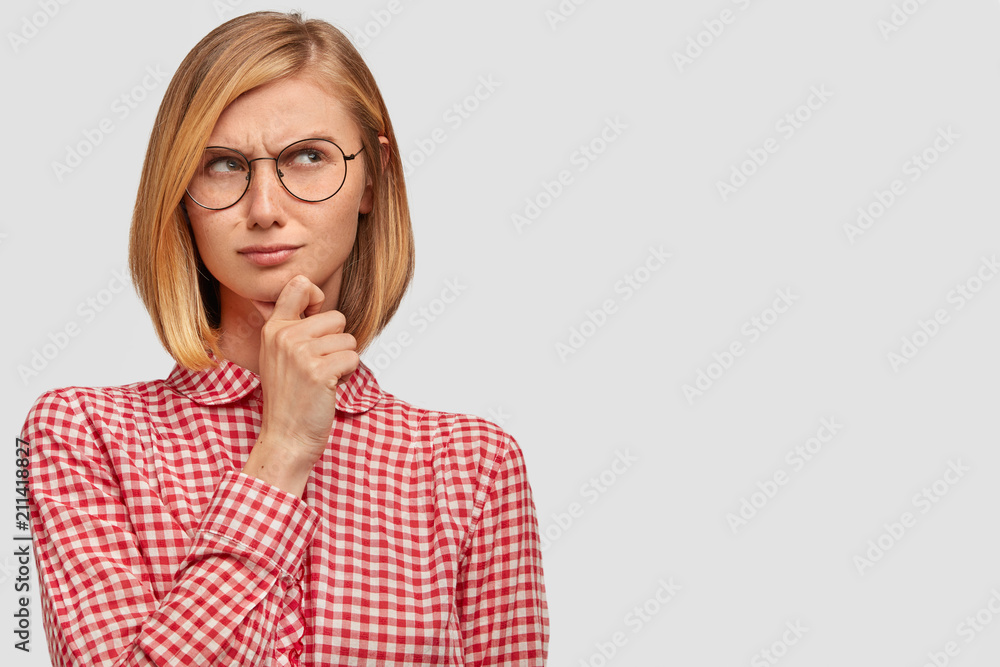 Pleasant looking thoughtful female tries to solve problem, keeps hand under chin, thinks about way out, dressed in checkered shirt, isolated over white background with copy space for your text