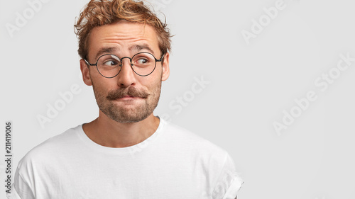 Doubtful puzzled bearded male with curious expression, curves lips and looks aside, concentrated on something, has intriguing look, stands against white background with copy space for your text