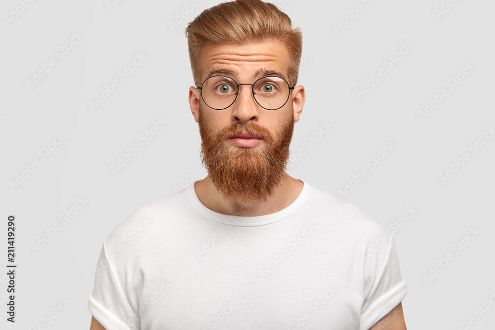 Obraz premium Photo of surprised young male with trendy haircut, has ginger beard and mustache, realizes that girlfriend didn`t come on date, stares with stupefied expression, isolated over white background