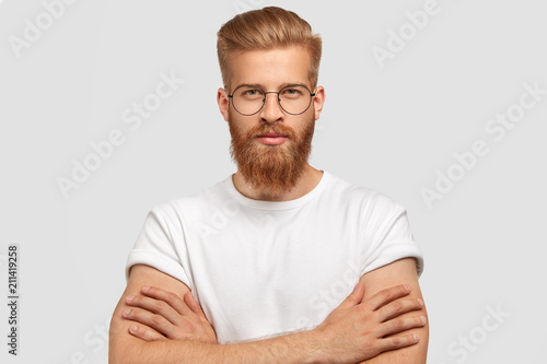 Studio shot of serious self assured male architect keeps hands crossed, has ginger thick beard and mustache, listens attentively client, stands against white background. Confident stylish hipster