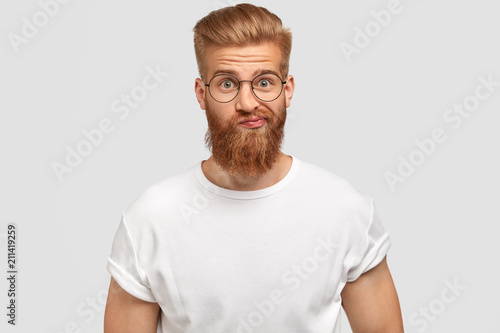 Handsome bearded male purses lips with hesitation, being surprised and puzzled to recieve unexpected news, dressed in casual white t shirt, poses against studio background. Trendy hipster indoor © wayhome.studio 