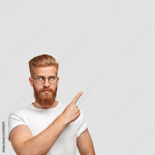 Serious bearded male with thick beard and mustache  looks aside  wears spectacles  indicates with index finger at upper right corner  show free space for your promotional text or advertisement
