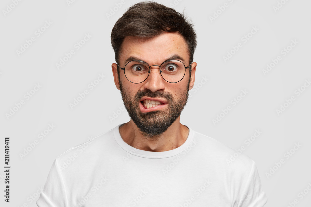 Fototapeta premium Displeased bearded male frowns face with displeasure, has irritated expression, raises eyebrows and clenches teeth, being discontent, argues with enemy, wears spectacles. Negative facial expressions
