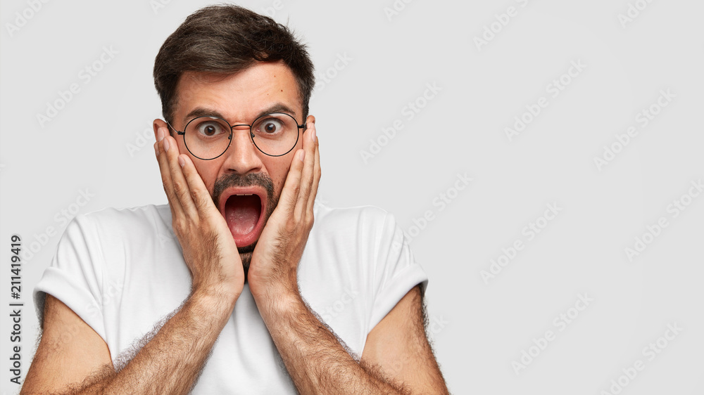 Horizontal shot of angry worried scared male with widely opened mouth,  touches cheeks with both palms, can`t believe in his failure, feels furious  and frustrated, stands over white wall, blank space Stock