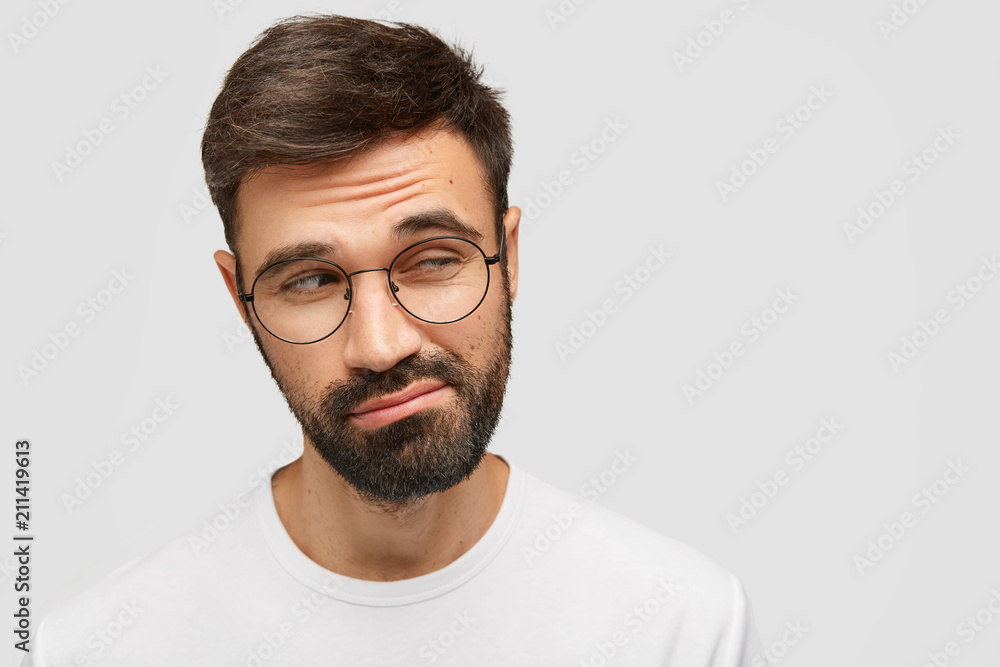 Clueless doubtful male with dark thick beard, looks with hesitation aside, raises eyebrows in bewilderment, has pensive expression, isolated over white background with copy space for advertisement - obrazy, fototapety, plakaty 