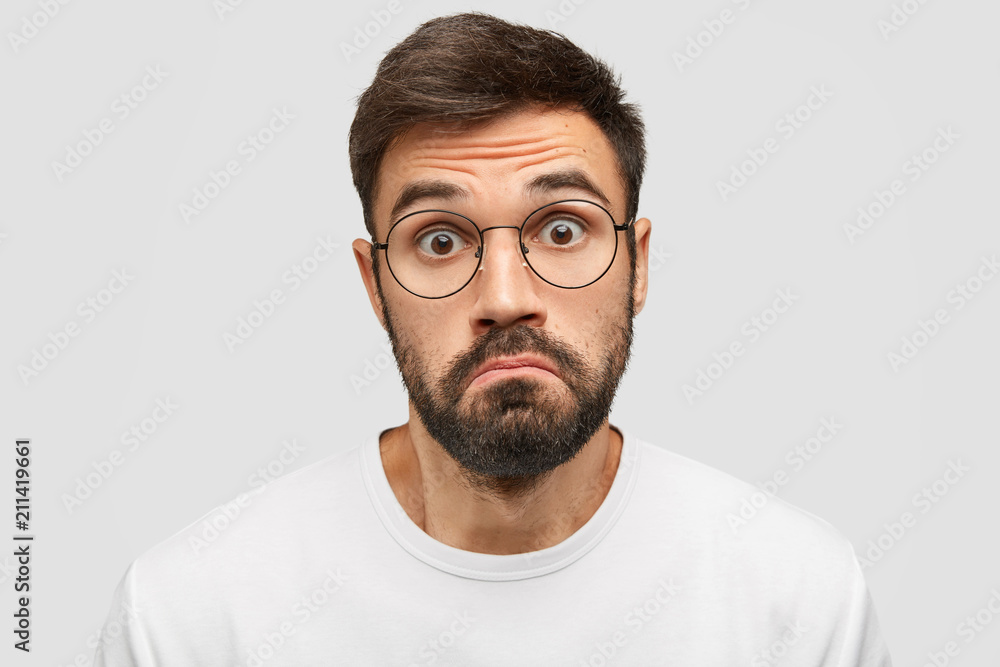 Doubtful bearded male purses lips with hesitation, stares at camera with scared expression, feels cluless while recieves unexpected offer from friend, isolated over white background. Facial expression Doubtful bearded male purses lips with hesitation, stares at camera with scared expression, feels cluless while recieves unexpected offer from friend, isolated over white background. Facial expression