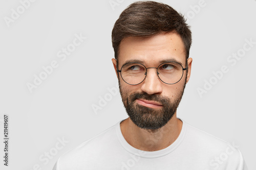 Headshot of attractive unshaven young male looks doubtfully, thoughtfullly aside, purses lips, has thick dark stubble, raises eyes upwards, focused on something, poses against white background photo