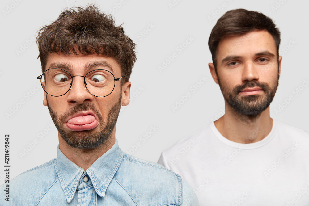 Funny male makes grimace, foolishes, crosses eyes and shows tongue, his bearded friend stands in background, has serious expression. Different emotions. Two young man companions play fool indoor Funny male makes grimace, foolishes, crosses eyes and shows tongue, his bearded friend stands in background, has serious expression. Different emotions. Two young man companions play fool indoor