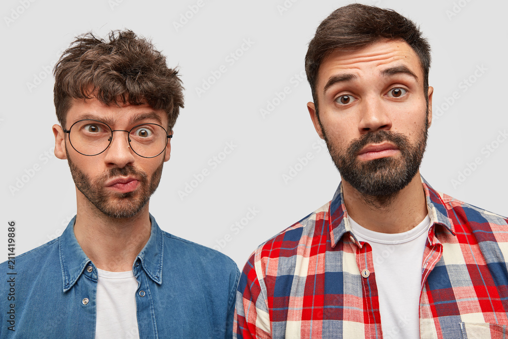 Two bearded male journalists have puzzled expressions, work on creating article, look at camera with bewilderment, dressed in fashionable shirts, stand next to each other, isolated over white wall Two bearded male journalists have puzzled expressions, work on creating article, look at camera with bewilderment, dressed in fashionable shirts, stand next to each other, isolated over white wall
