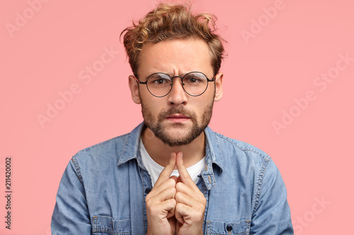 Headshot of serious intelligent young male proffesor listens something with attentive look, has scrupulous gaze directly into camera, dressed in fashionable outfit, isolated over pink background