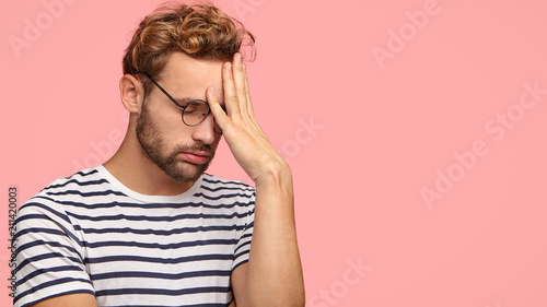 Attractive tired overworked male entrepreneur sighs with tiredness, keeps hand on forehead, suffers from headache, hangs head, stands in profile against pink background. Omg, I am fed up of everything photo
