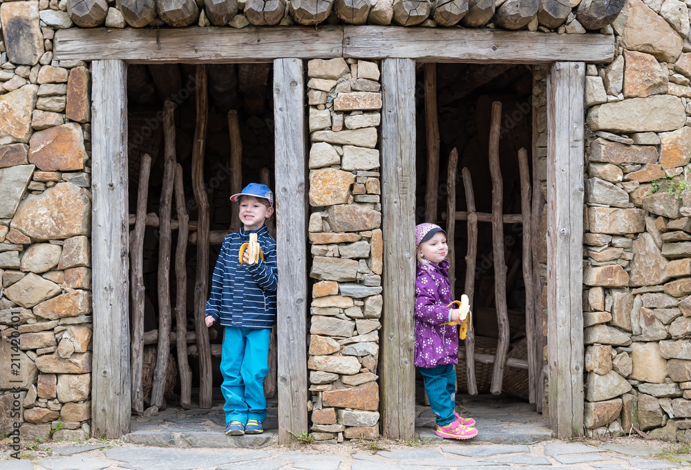 Two children are standing in different doors of an old stone house having banana for a snack