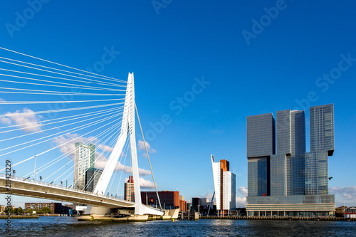 Cityscape of the Dutch city Rotterdam with high rise buildings in the financial district and port area with the Erasmus bridge seen from the water against a blue sky with fluffy clouds