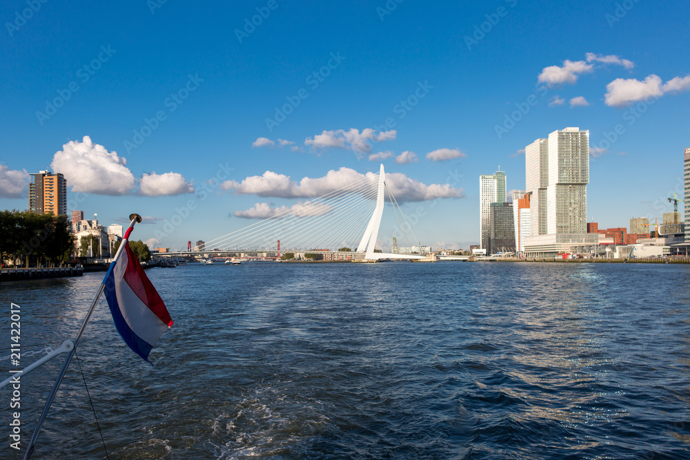 Cityscape of Rotterdam with modern high rise buildings and the Erasmus bridge in the background and the Dutch national red, white and blue flag in the foreground against a clear blue sky with clouds