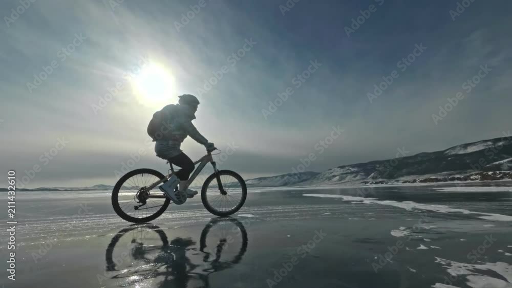 Woman is riding bicycle on the ice. The girl is dressed in a silvery down jacket, cycling backpack and helmet. Ice of the frozen Lake Baikal. The tires on the bicycle are covered with special spikes Woman is riding bicycle on the ice. The girl is dressed in a silvery down jacket, cycling backpack and helmet. Ice of the frozen Lake Baikal. The tires on the bicycle are covered with special spikes