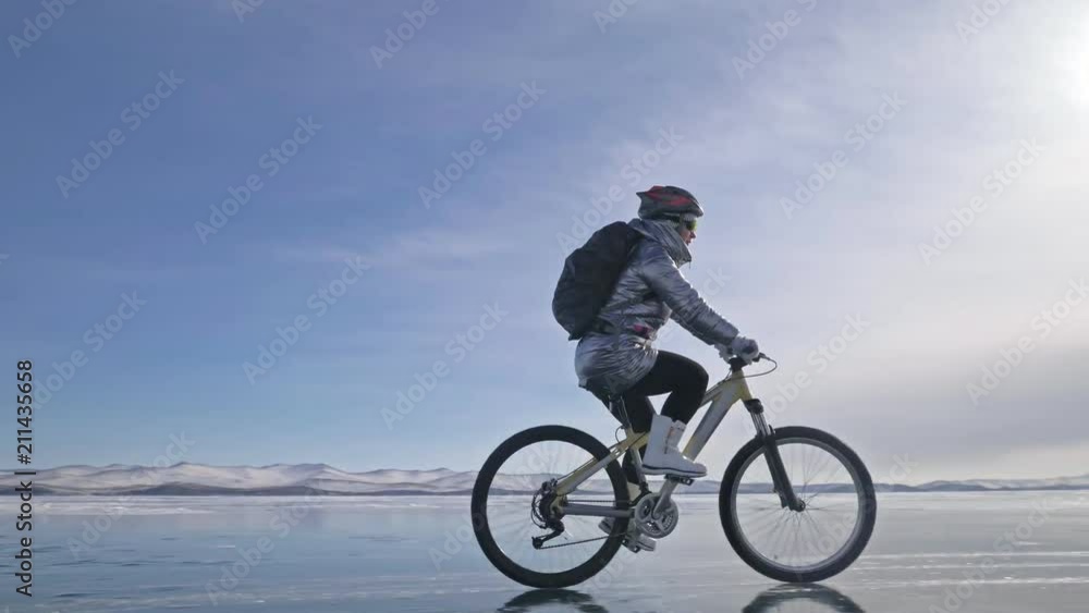Woman is riding bicycle on the ice. The girl is dressed in a silvery down jacket, cycling backpack and helmet. Ice of the frozen Lake Baikal. The tires on the bicycle are covered with special spikes Woman is riding bicycle on the ice. The girl is dressed in a silvery down jacket, cycling backpack and helmet. Ice of the frozen Lake Baikal. The tires on the bicycle are covered with special spikes