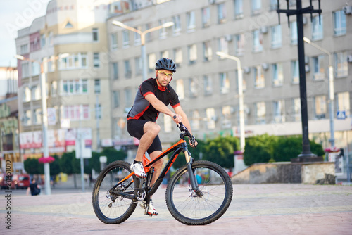 Athletic strong man cyclist posing in professional cycling garment on bicycle in front of city building. Sportsman exercising outdoors, rest after working day. Concept of healthy sporty lifestyle