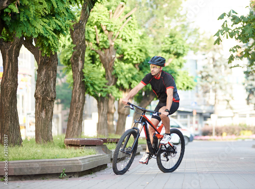 Athletic guy bicyclist in cycling clothes and protective gear riding on bicycle along empty city streets, green trees around. Sportsman performing morning outdoor exercise and training. © anatoliy_gleb