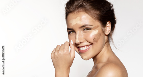 Waist up portrait of cheerful lovable brunette applying tonal basis different shades for perfect natural maquillage. Beauty treatment concept. Isolated on grey background