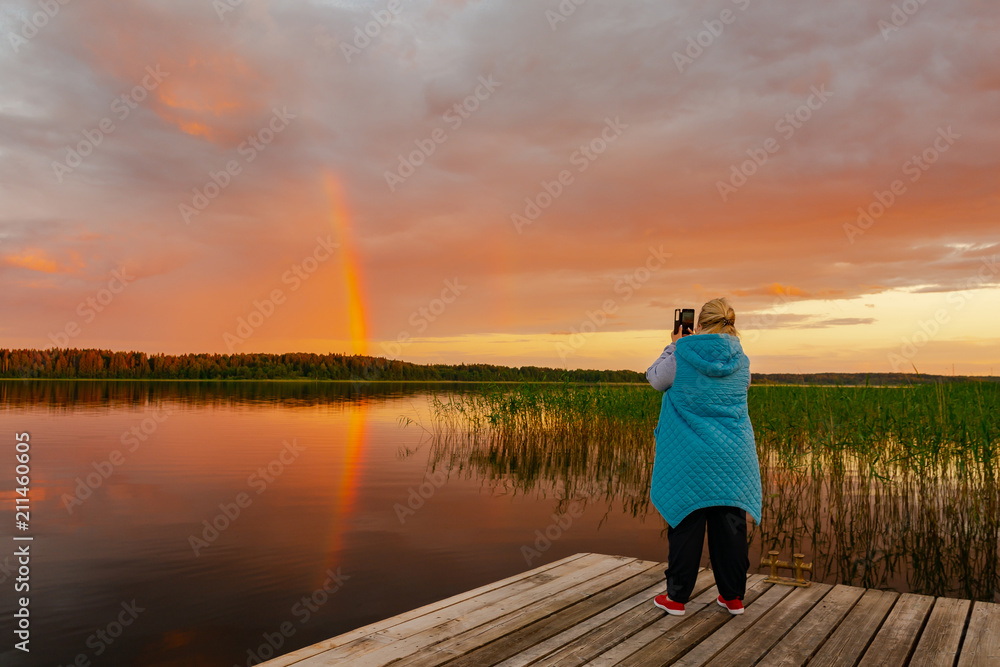 Summer evening. A woman stands on the dock and takes a photo of the rainbow.
