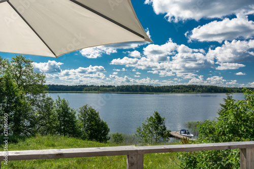 Summer landscape on the lake. Left umbrella. In the foreground of the railing. Berth with a boat downstairs. A forest in the distance.