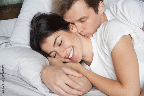 Romantic young husband waking up beautiful wife by kiss in neck, loving millennial couple sleeping hugging each other in cozy bed a home, spouses relaxing lying and embracing during lazy morning