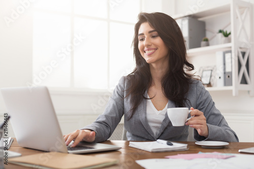 Smiling businesswoman working on laptop and drinking coffee at office