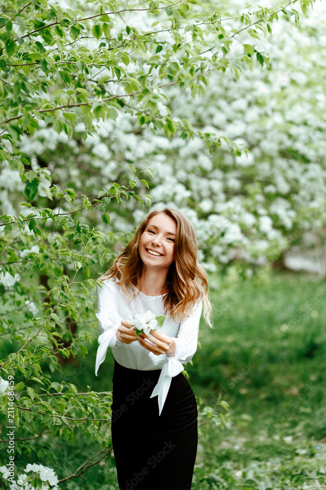 Cute spring portrait. Young business woman in a business office suit on the background of Apple trees in bloom. Girl holding a white flower.