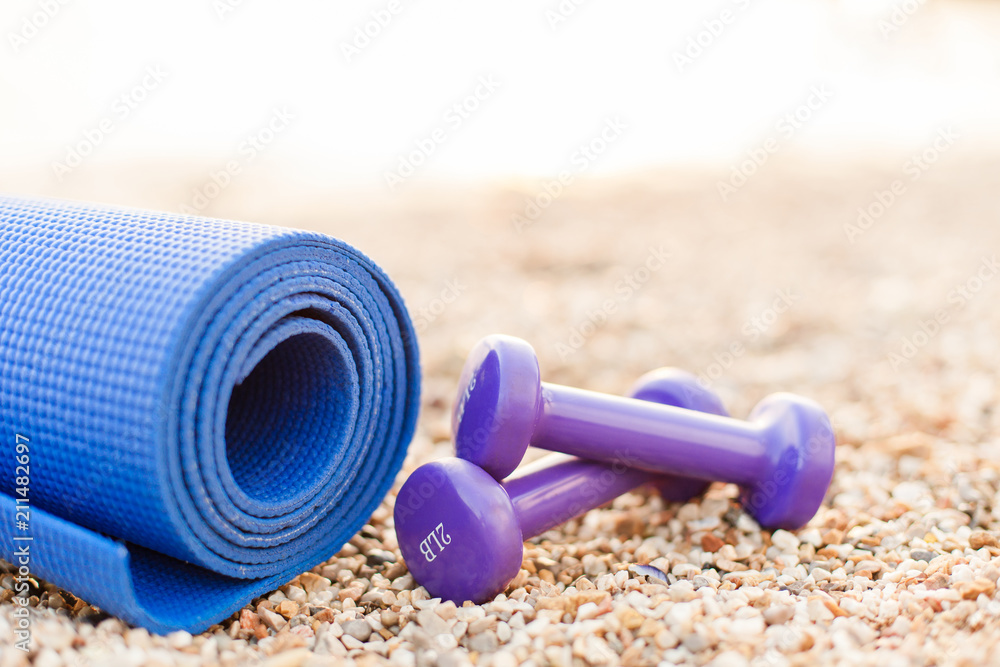 Two purple dumbbells and blue fitness yoga mat on stone and pebble sea beach. Sport background. Concept of healthy lifestyle and traveling. Sport female equipment. Copy space. Two purple dumbbells and blue fitness yoga mat on stone and pebble sea beach. Sport background. Concept of healthy lifestyle and traveling. Sport female equipment. Copy space.