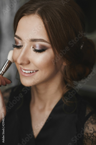 Makeup artist applying makeup on face of a beautiful and cheerful brunette model girl with charming smile and closed eyes  in black peignoir sitting in interior