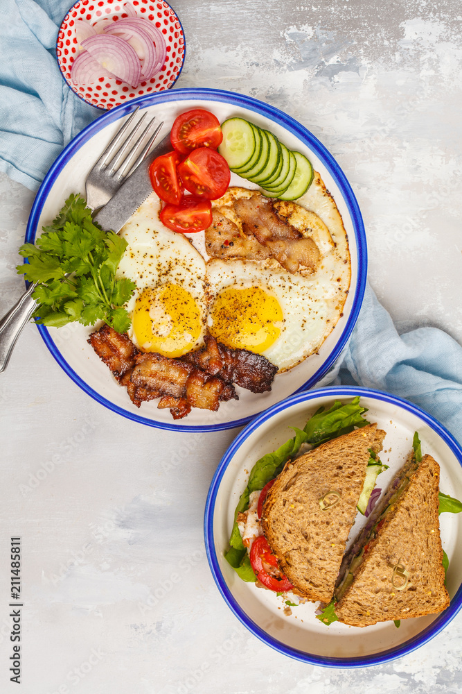 Fried eggs with bacon and a sandwich with meat, cheese and vegetables on white background, top view.
