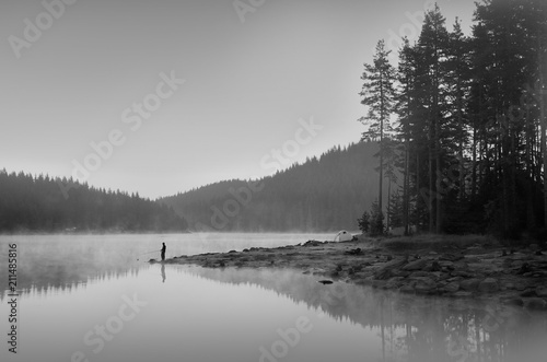 Magic reflections in the water of the dam. Sunrise in Rhodope mountain, Bulgaria. A man is fishing early in the morning.