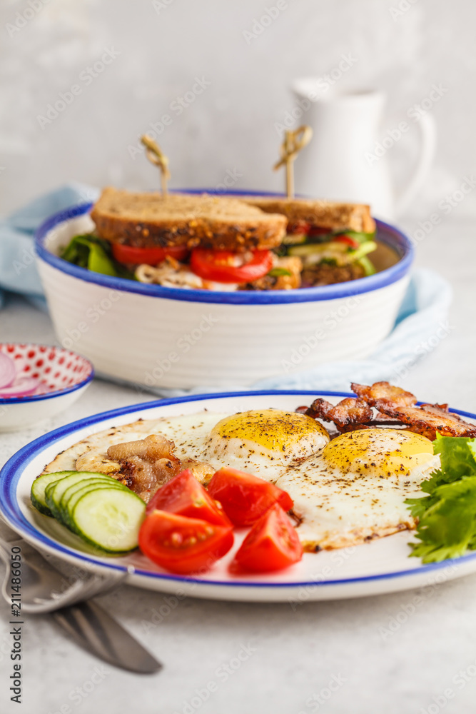 Fried eggs with bacon and a sandwich with meat, cheese and vegetables on white background. Delicious hearty breakfast. Fried eggs with bacon and a sandwich with meat, cheese and vegetables on white background. Delicious hearty breakfast.